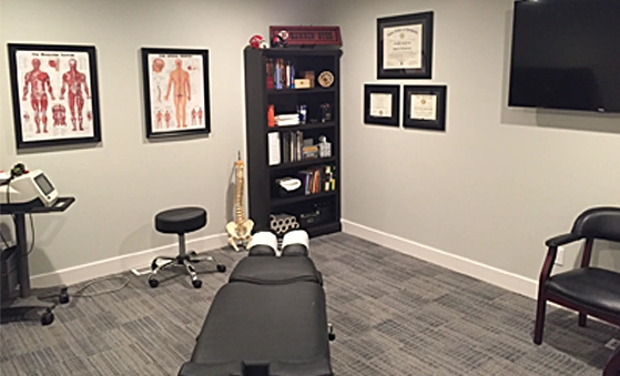 Chiropractic office with an adjustment table, anatomical charts on the wall, a black bookshelf with books and spine model, framed certificates, and a mounted TV