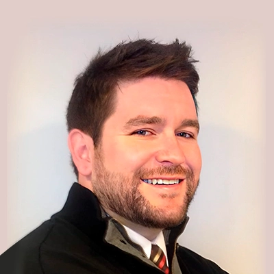 Dr. Brad Anderson wearing a black jacket over a collared shirt with a striped tie, shown from the shoulders up against a light-colored background