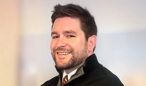Doctor Brad Anderson wearing a black jacket over a collared shirt with a striped tie, standing against a softly blurred background